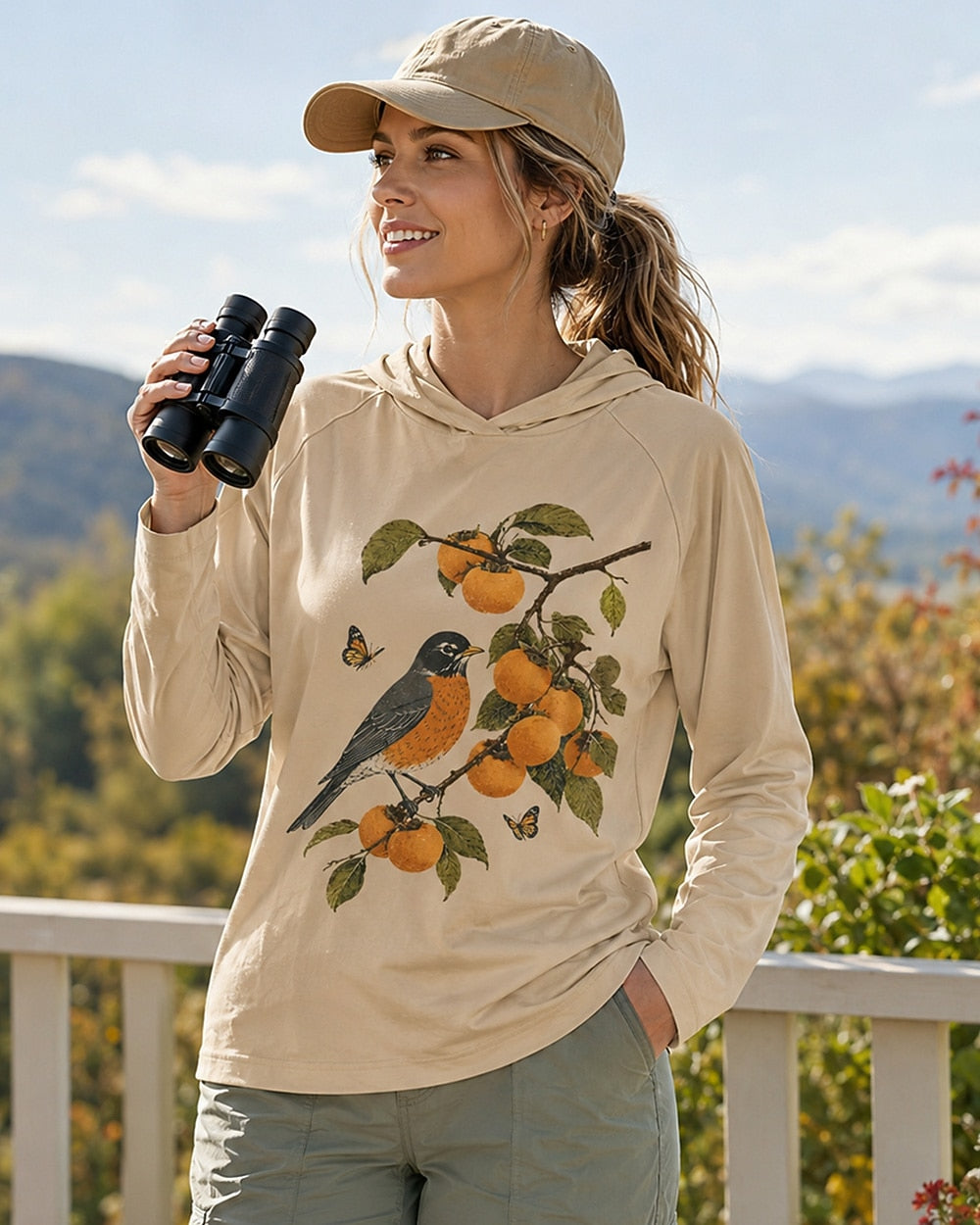 Woman holding binoculars outdoors in a light yellow bluebird and orange tree sun hoodie.