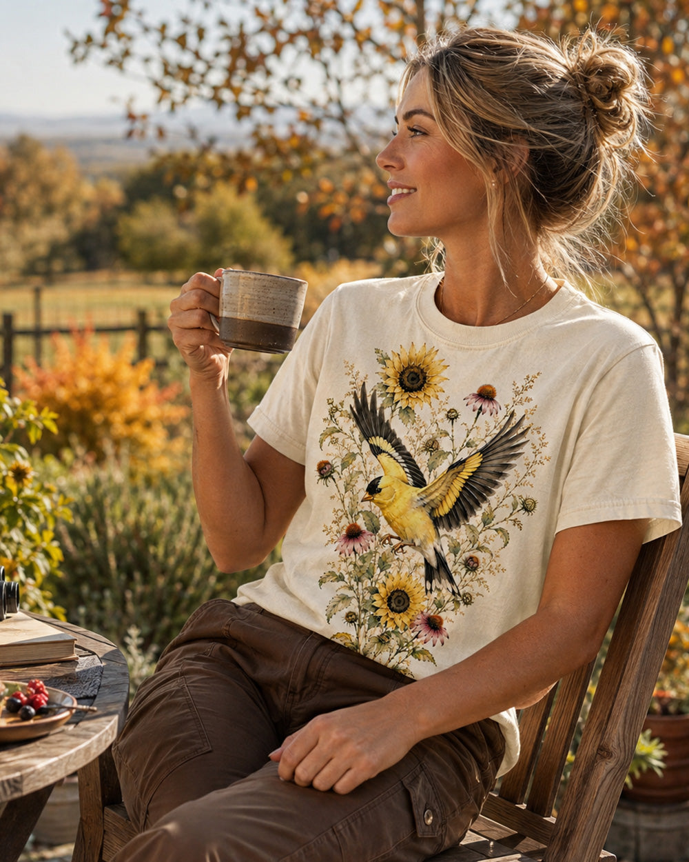 Woman wearing a off white cotton T-shirt with goldfinches in flight among sunflowers outdoors.