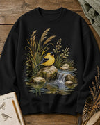 Women's black crewneck sweatshirt with a goldfinch beside a rocky waterfall, front view.