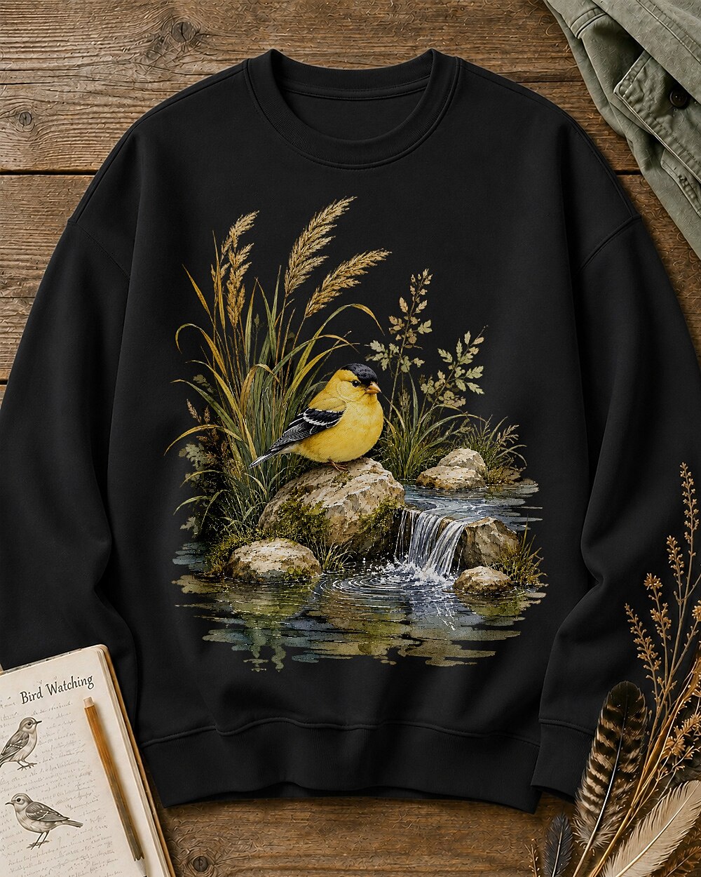 Women's black crewneck sweatshirt with a goldfinch beside a rocky waterfall, front view.