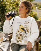 Woman wearing a white crewneck sweatshirt with a goldfinch among yellow flowers outdoors.
