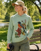 Woman wearing a green sun hoodie with a robin perched on a red berry branch outdoors.