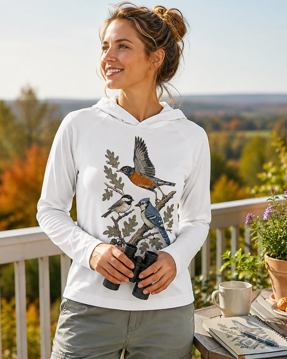 Woman wearing a white sun hoodie with a flying robin and blue jay among branches outdoors.