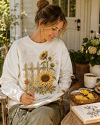 Woman wearing a white crewneck sweatshirt with sunflowers beside a picket fence outdoors.