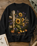 Women's black crewneck sweatshirt with sunflowers beside a rustic wooden fence, front view.